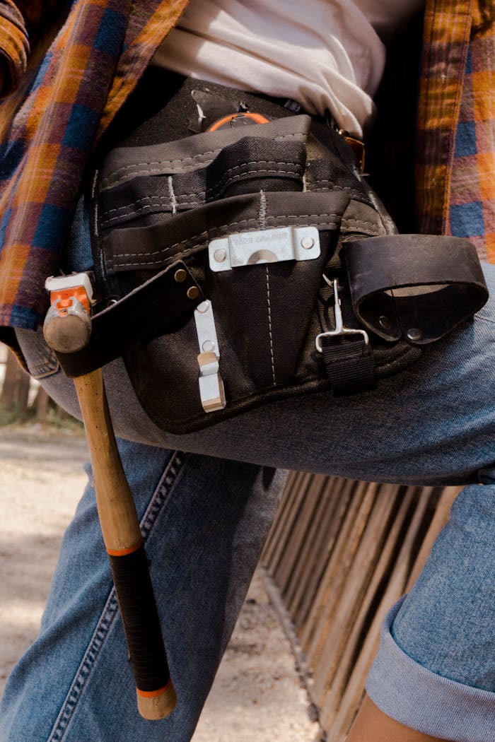 Close-up of a utility belt filled with tools on a person wearing jeans and a flannel shirt outdoors.